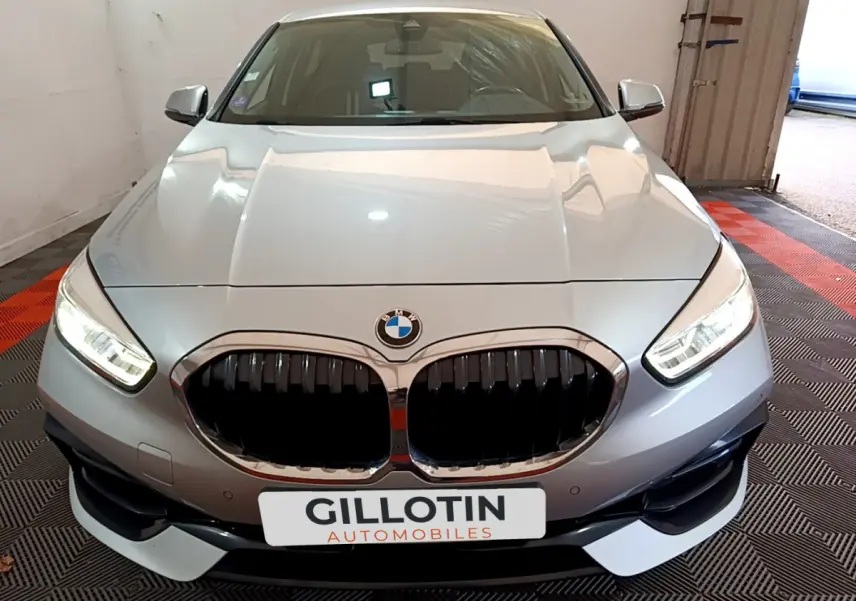 Vue avant d'une BMW Série 1 gris clair avec calandre noire et phares LED allumés en intérieur showroom.