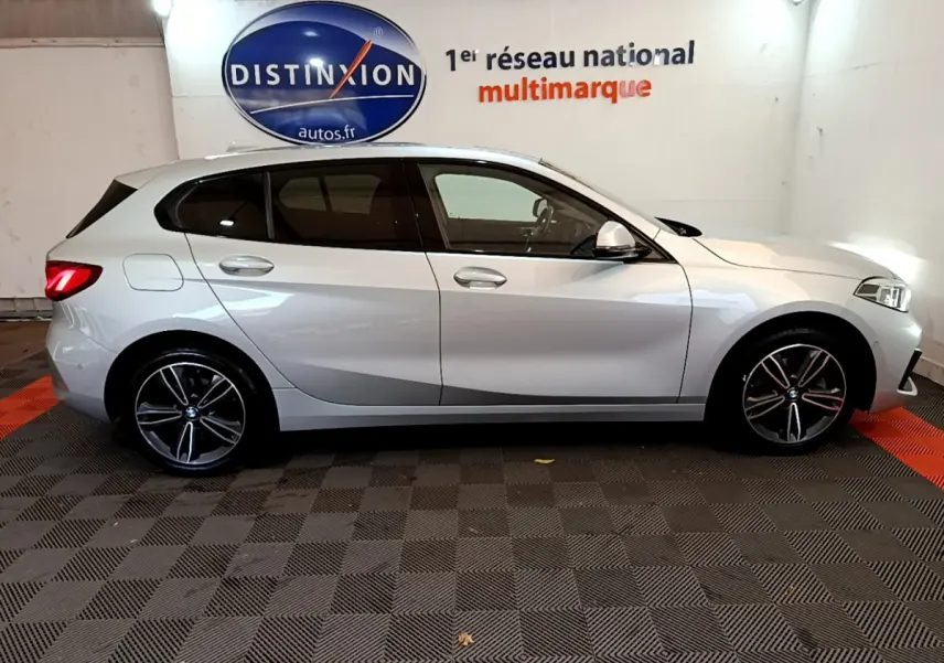 BMW Série 1 gris clair vue de profil côté gauche, avec jantes alliage et feux arrière allumés dans un showroom.