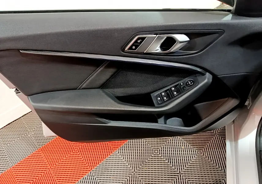 Porte côté conducteur ouverte d'une BMW Série 1 gris clair, montrant les commandes électriques des vitres et la poignée intérieure noire.