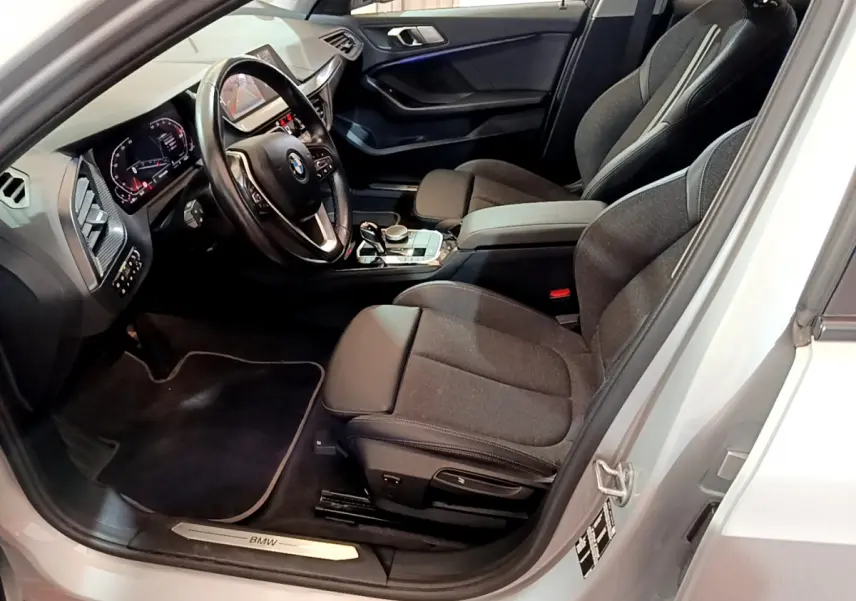 Intérieur avant droit d'une BMW Série 1 gris clair, montrant sièges tissu, volant cuir multifonctions et tableau de bord digital.