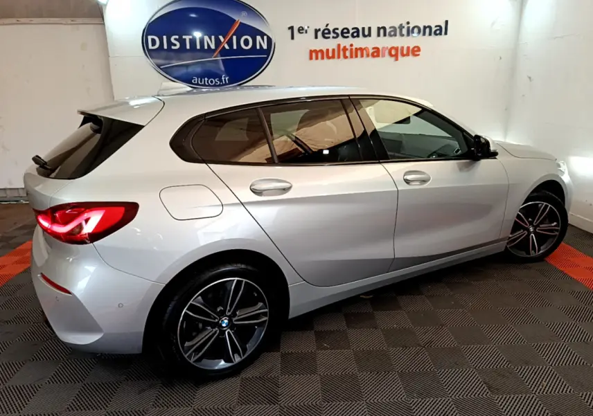 BMW Série 1 gris clair vue de profil côté droit, avec jantes alliage et feux arrière allumés en intérieur showroom.