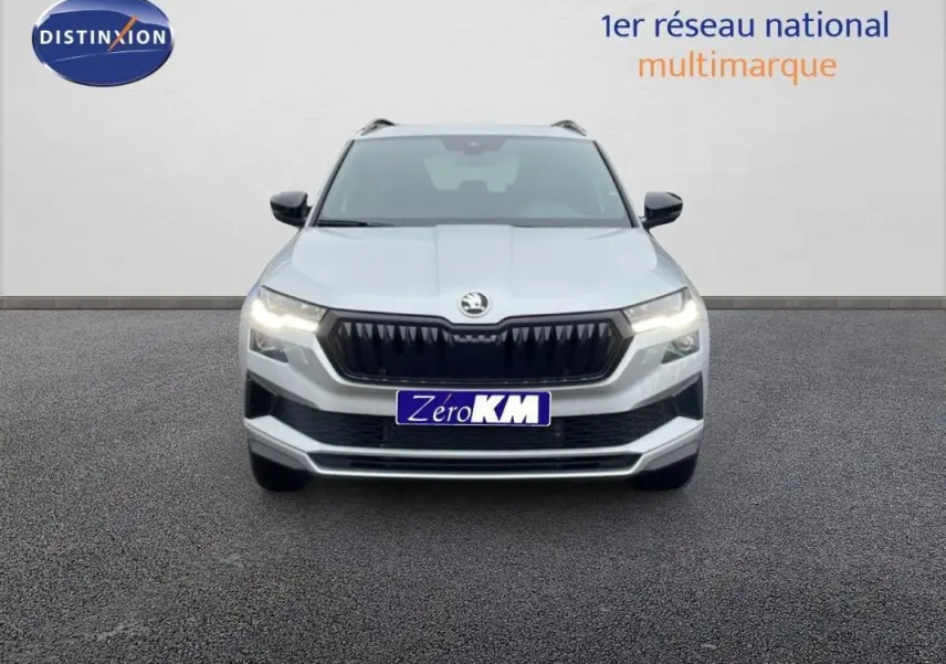 Vue frontale d'un Skoda Karoq argent 2025 avec calandre noire, phares allumés et barres de toit visibles.