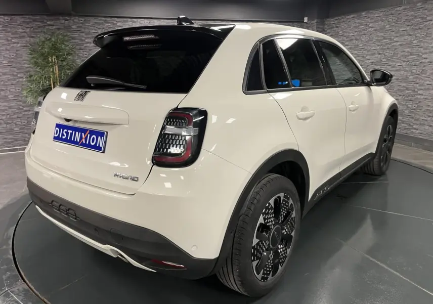 Vue 3/4 arrière droite d'une FIAT 600 blanche hybride 2024 avec jantes alliage noires et toit noir brillant.