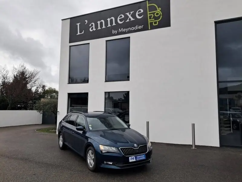 Skoda Superb Combi bleu métal 2018, vue 3/4 avant droit devant un bâtiment blanc avec enseigne L'annexe.
