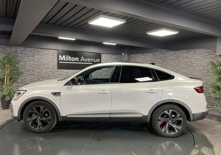 Profil côté gauche d'un Renault Arkana blanc perle 2022 R.S. Line avec jantes noires et accents rouges en intérieur showroom.