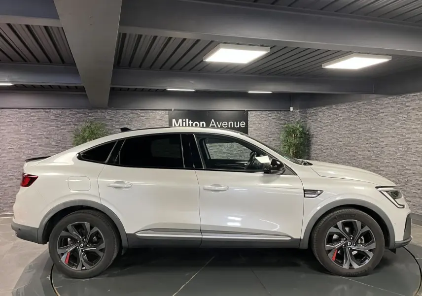 Profil côté gauche du Renault Arkana blanc perle 2022 avec jantes noires et toit noir brillant en intérieur showroom.