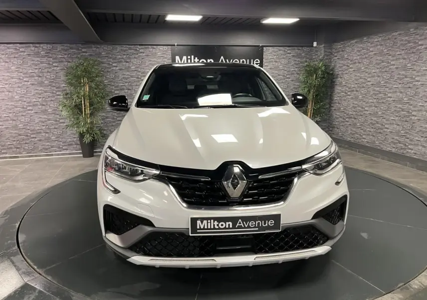 Vue frontale d'un Renault Arkana blanc perle avec calandre noire et logo Renault au centre, en showroom.