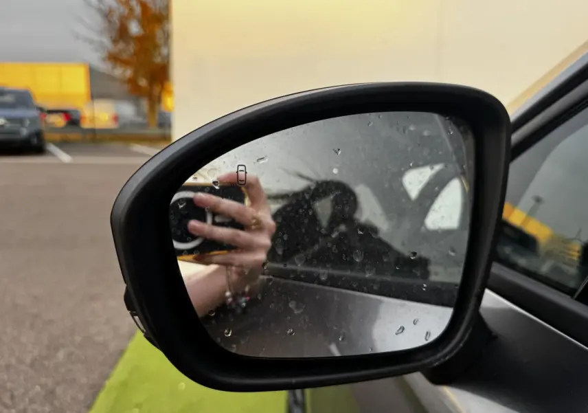 Rétroviseur extérieur droit noir brillant de Renault Clio gris schiste, avec traces d'eau et reflet de main tenant un appareil photo.