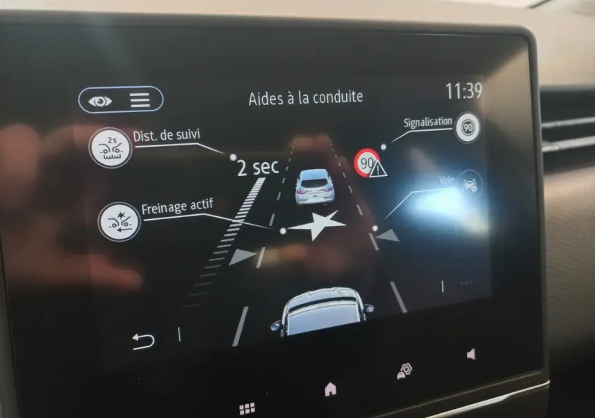 Écran tactile intérieur montrant l’aide à la conduite avec affichage de distance de suivi et freinage actif sur Renault Clio blanche.