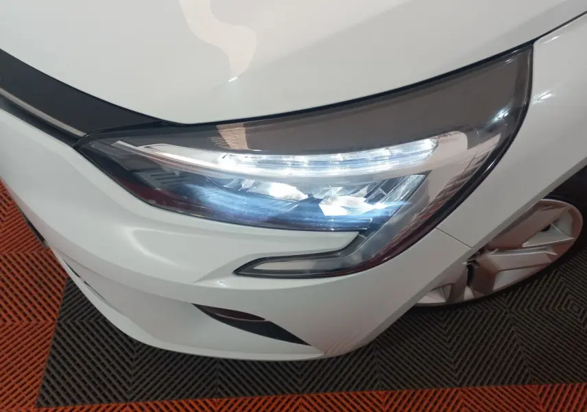 Gros plan sur le phare avant droit allumé d’une Renault Clio Business blanche, avec partie de la roue visible.