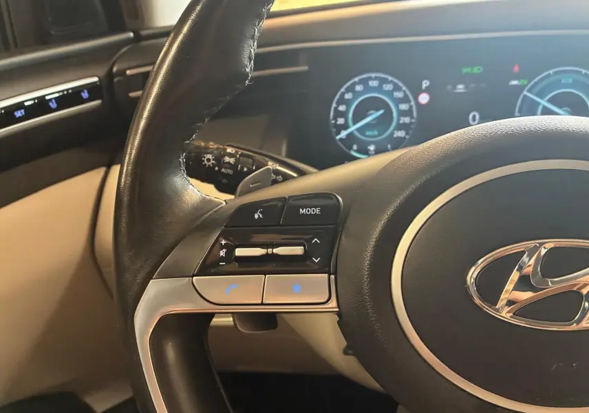 Vue rapprochée du volant noir et beige du Hyundai Tucson 2022, avec commandes intégrées et tableau de bord numérique en arrière-plan.