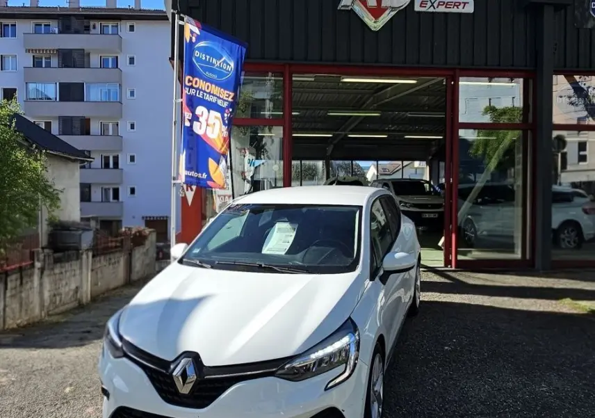 Renault Clio blanc glacier vue 3/4 avant droit devant un garage AD Expert avec une affiche promotionnelle bleue.