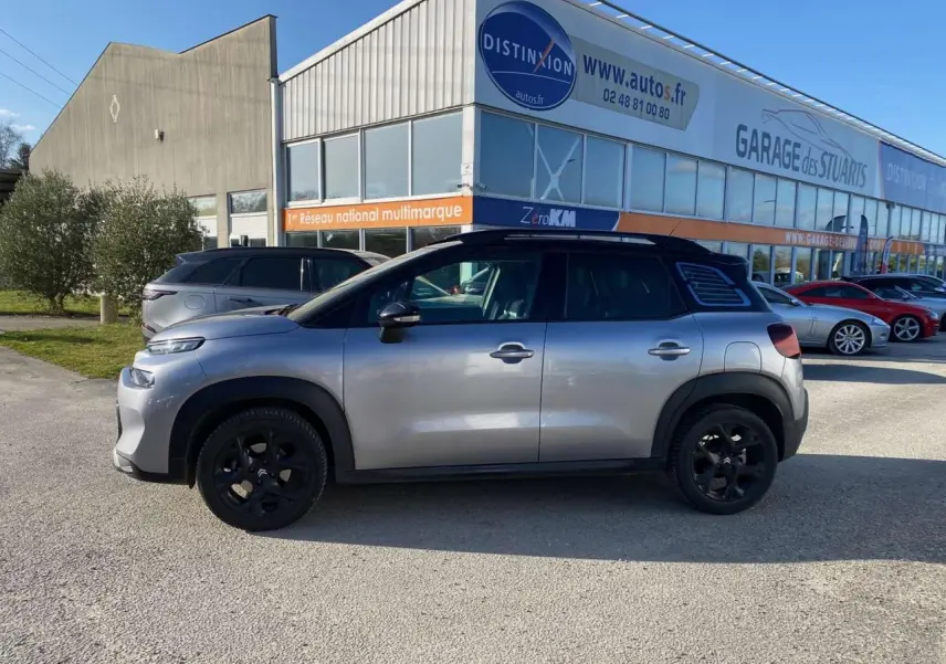 Vue de profil côté gauche d'un Citroën C3 Aircross gris foncé 2024 avec jantes noires, stationné devant un garage.