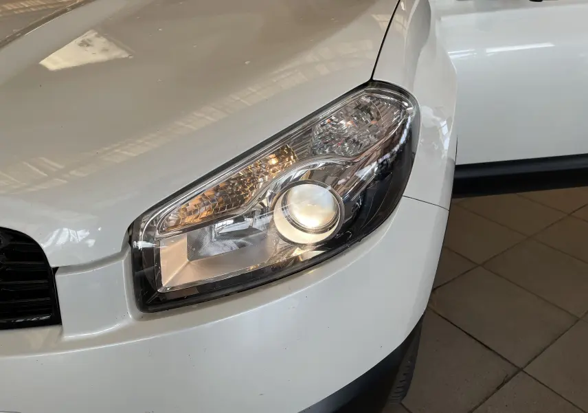 Gros plan sur le phare avant droit du Nissan Qashqai blanc de 2013, mettant en valeur son design moderne et épuré.