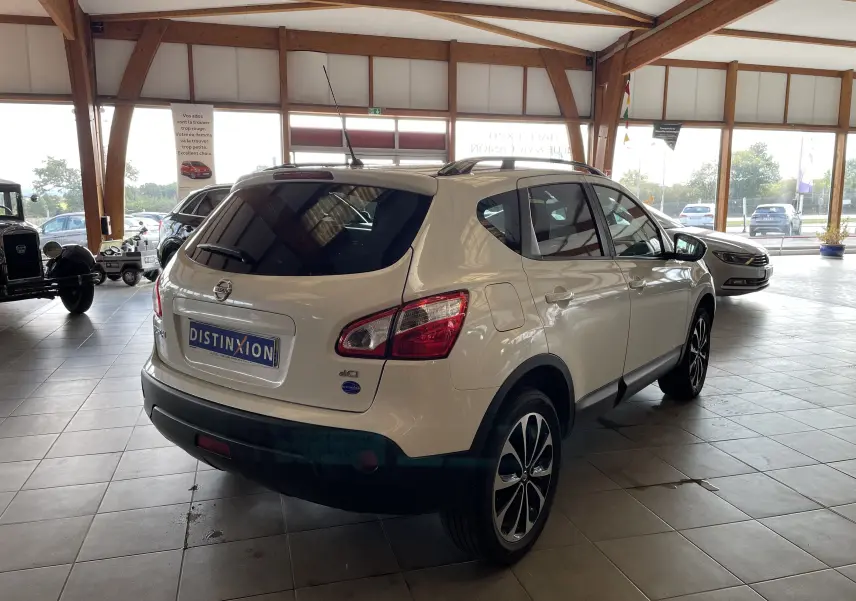 Vue 3/4 arrière droite d'un Nissan Qashqai blanc 2013 en showroom, avec jantes alliage et vitres teintées.