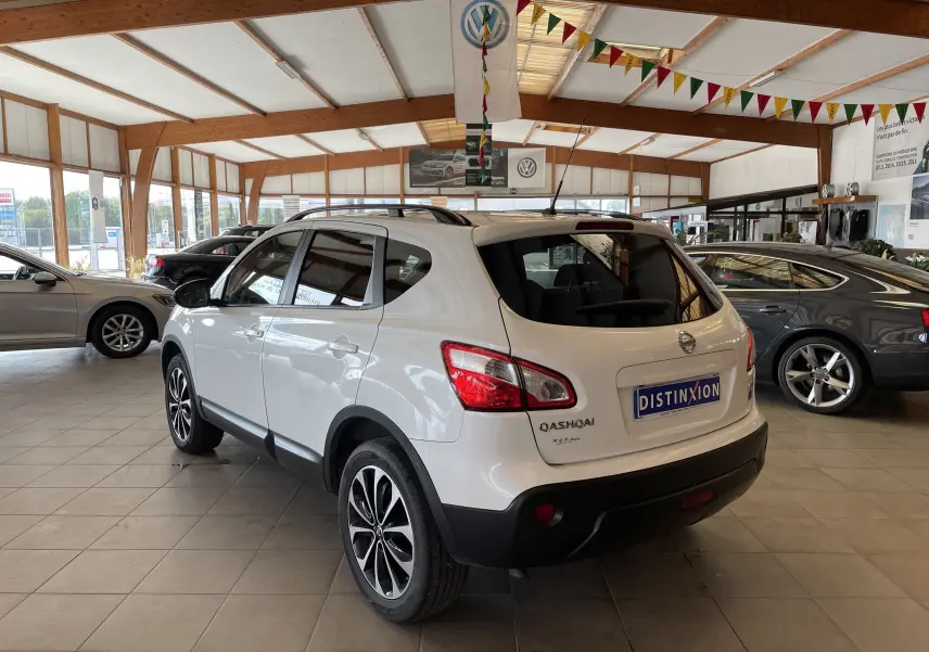 Vue 3/4 arrière droite d'un Nissan Qashqai blanc 2013 en showroom, avec jantes alliage et toit ouvrant visible.
