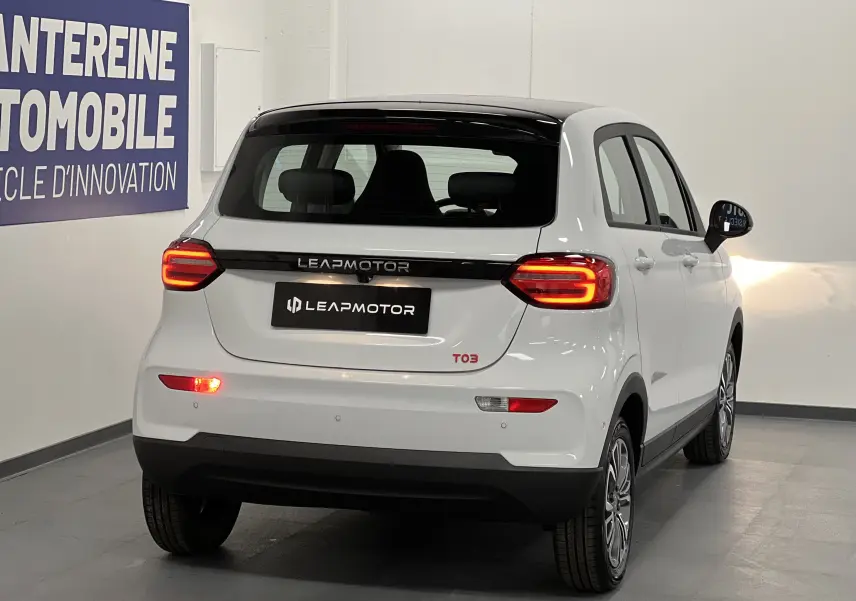 Vue 3/4 arrière droite d'une Leapmotor T03 blanche avec feux arrière allumés dans un showroom intérieur.