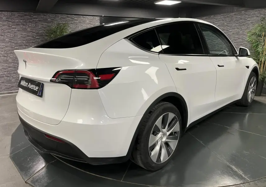 Tesla Model Y blanc nacré vu en 3/4 arrière droit dans un showroom avec jantes alu et toit panoramique noir.