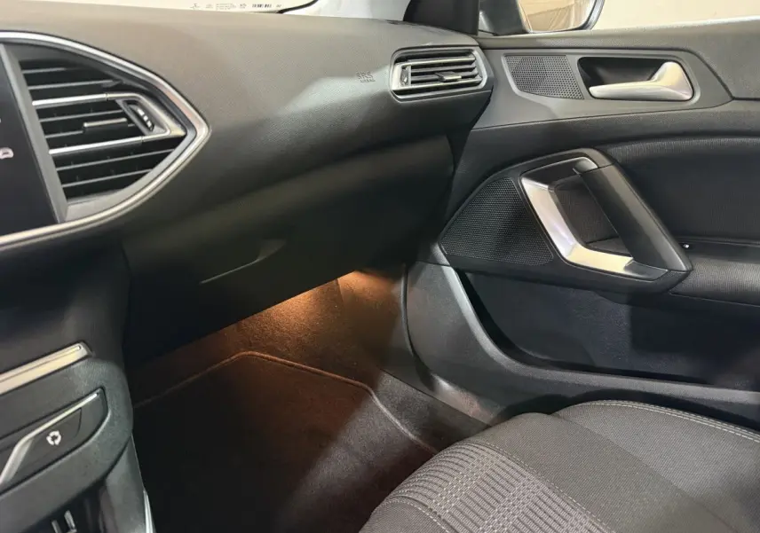 Vue intérieure côté passager de la Peugeot 308 noire 2020, montrant tableau de bord et porte avec éclairage d'ambiance au sol.