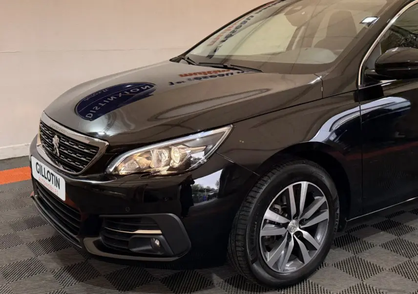 Vue 3/4 avant droit d'une Peugeot 308 noire avec phares allumés et jantes alliage visibles.