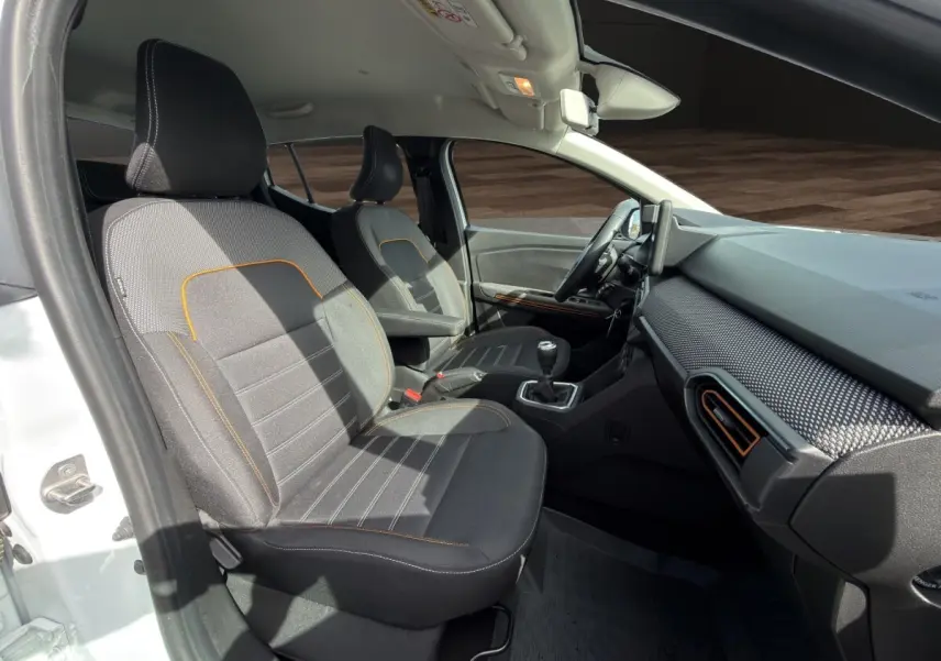 Vue intérieure côté conducteur de la Dacia Sandero Stepway blanche, sièges tissu gris avec surpiqûres orange et tableau de bord moderne.