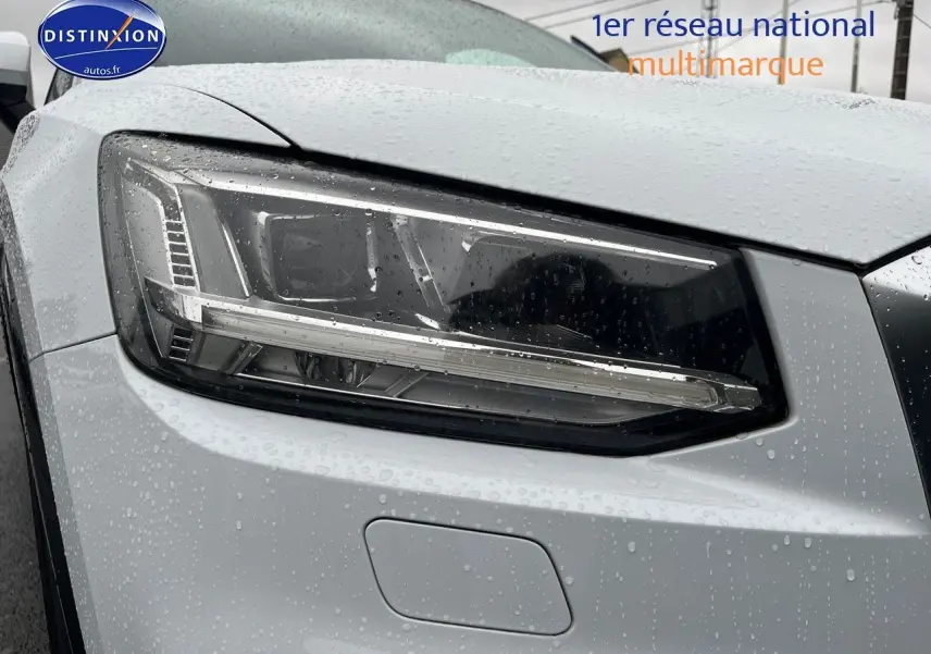 Gros plan sur le phare avant gauche d'un Audi Q2 blanc Ibis sous la pluie, mettant en valeur son design LED.