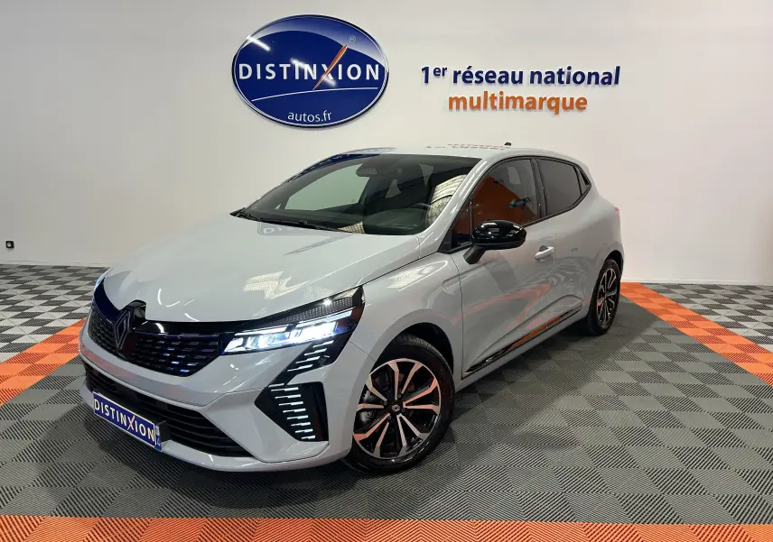 Renault Clio V E-Tech gris Rafale métal, vue 3/4 avant droit, phares LED allumés et jantes noires bicolores.