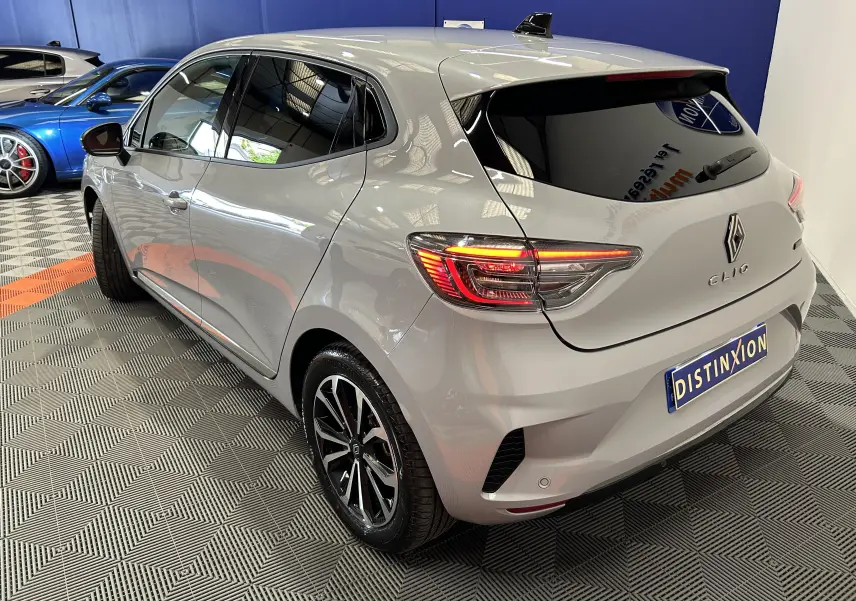 Vue 3/4 arrière droite d’une Renault Clio V E-Tech gris Rafale métallisé avec feux arrière allumés en intérieur showroom.