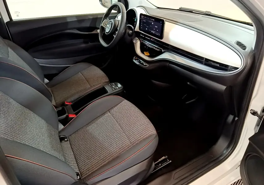 Intérieur avant droit de la Fiat 500 électrique blanche 2022, sièges tissu gris et tableau de bord avec écran tactile central.