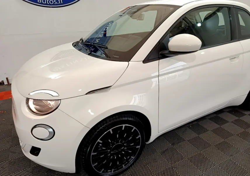Vue 3/4 avant droit d'une Fiat 500 électrique blanche avec jantes noires et phares allumés.