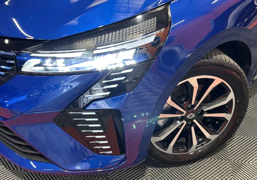 Gros plan sur l'avant droit bleu Iron Metal de la Renault Clio V avec phares LED allumés et jante alliage distinctive.