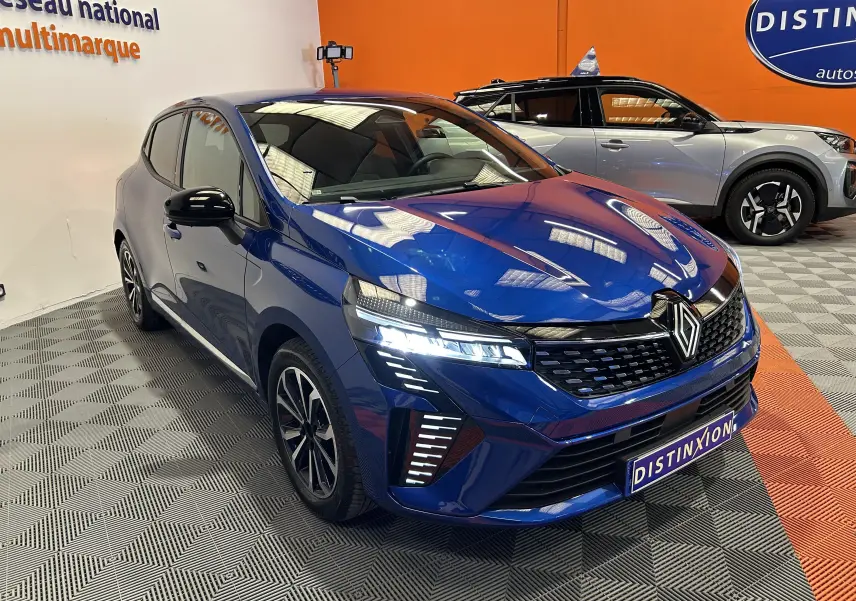 Vue 3/4 avant d'une Renault Clio V bleu Iron Metal avec phares LED allumés dans un showroom.