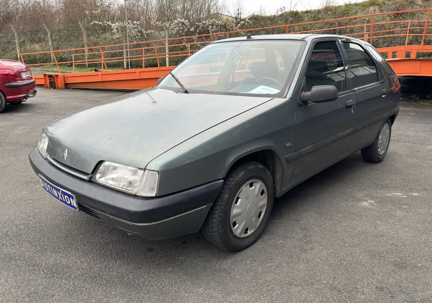 Citroën ZX 1.6i gris vue 3/4 avant droit, avec jantes pleines et rétroviseurs noirs sur parking extérieur.