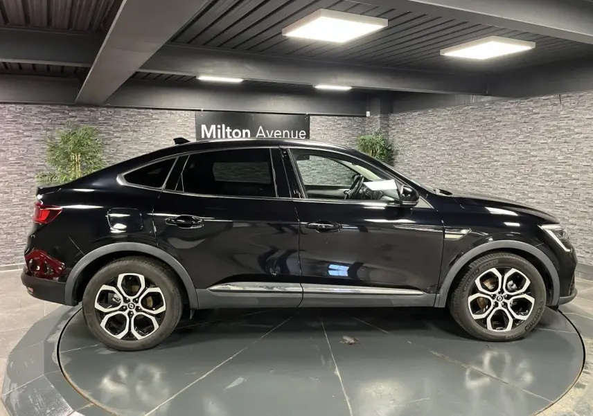 Profil côté gauche du Renault Arkana noir métallisé 2021 avec jantes alliage et lignes fluides dans un showroom intérieur.