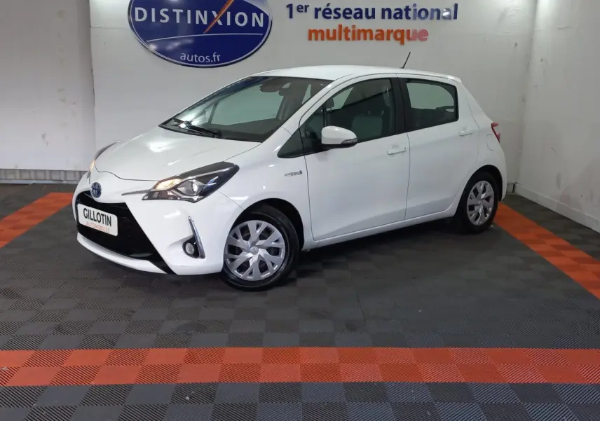 Toyota Yaris blanche vue de profil côté gauche en intérieur, avec phares allumés et logo hybride visible sur l'aile avant.