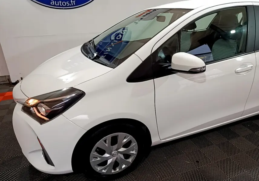 Toyota Yaris blanche vue 3/4 avant droit, phare allumé et rétroviseur avec clignotant intégré.
