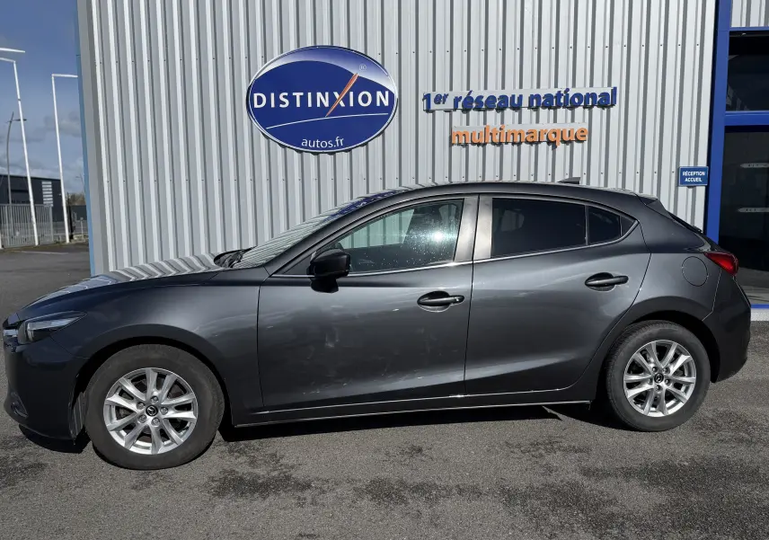 Vue de profil côté gauche d'une Mazda3 gris foncé stationnée devant un bâtiment avec un panneau Distinxion.
