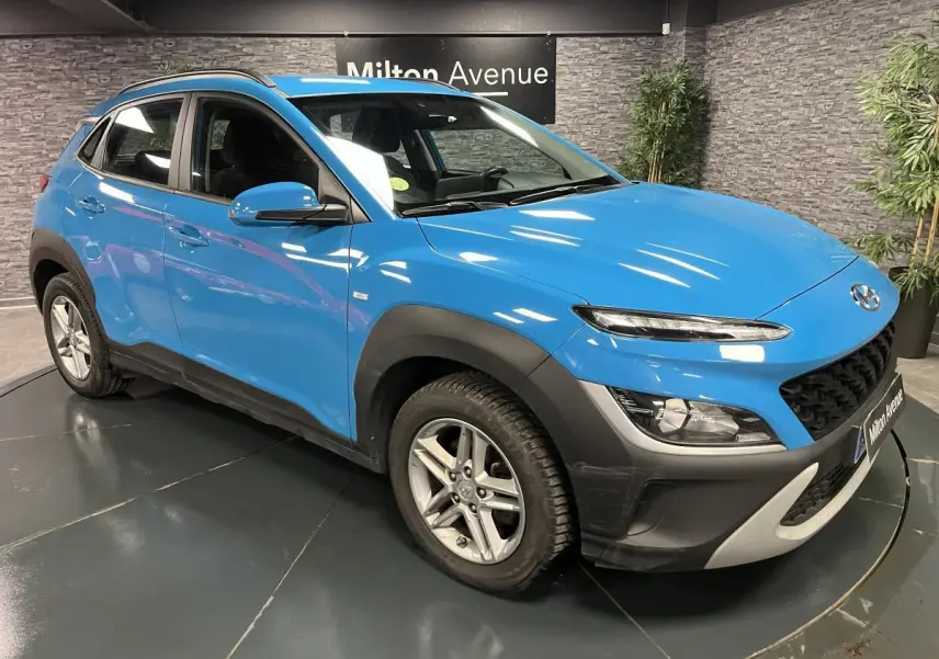 Hyundai Kona bleu 3/4 avant droit avec protections noires et jantes alliage 16 pouces en intérieur showroom.