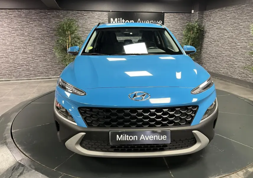 Vue avant d'un Hyundai Kona bleu avec calandre noire alvéolée et pare-chocs gris dans un showroom intérieur.