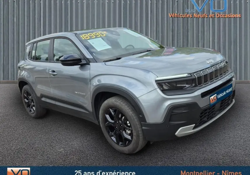 Jeep Avenger gris foncé en 3/4 avant droit, avec jantes noires et calandre distinctive, stationné devant un mur métallique.