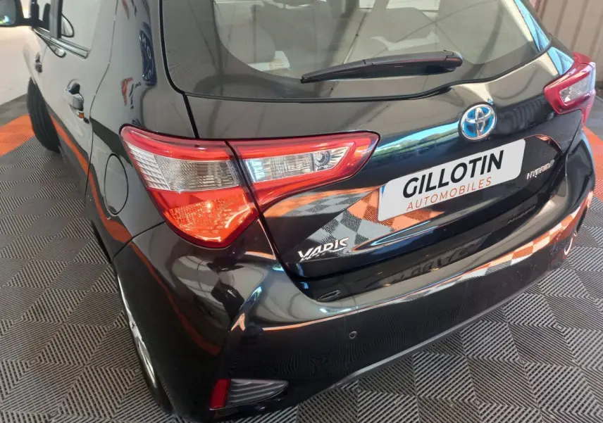 Vue 3/4 arrière gauche d'une Toyota Yaris noire hybride avec feux arrière distinctifs et logo visible.