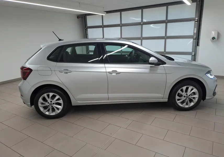 Vue de profil côté gauche d'une Volkswagen Polo 2025 en peinture reflet d'argent, avec feux avant allumés en intérieur showroom.