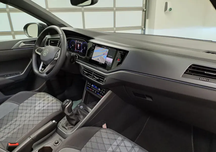Vue intérieure côté conducteur du Volkswagen Taigo 2025, tableau de bord noir avec écran tactile et sièges tissu à motifs gris.