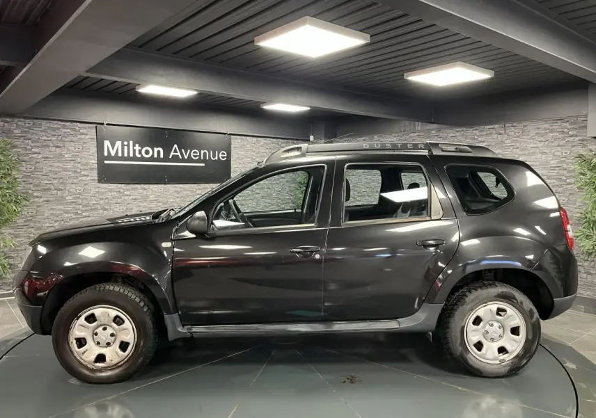 Profil côté gauche d'un Dacia Duster noir 2016 avec barres de toit et jantes acier en showroom.