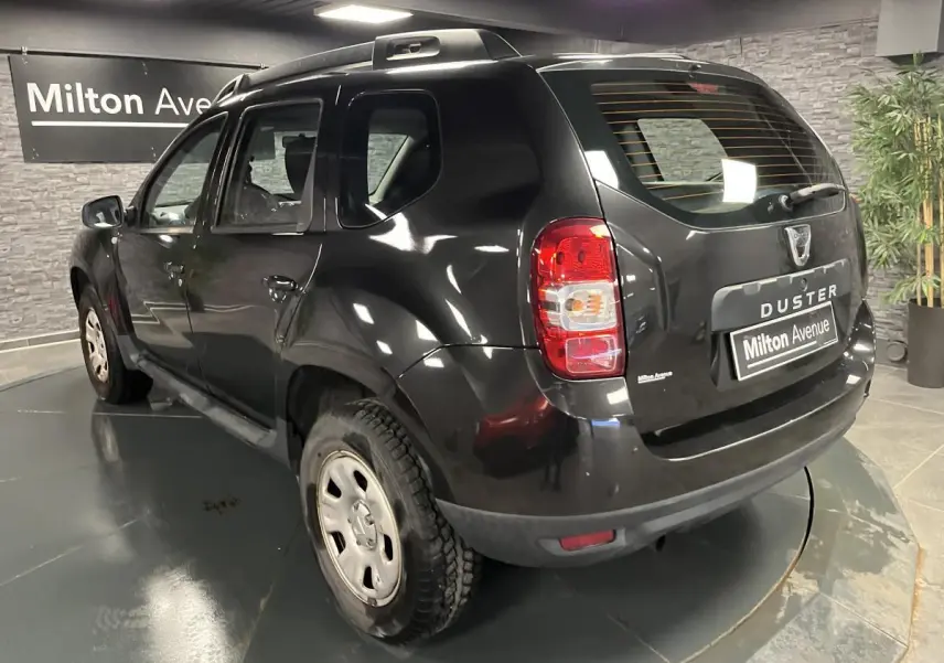 Vue 3/4 arrière gauche d'un Dacia Duster noir 2016 avec barres de toit et jantes acier en intérieur showroom.
