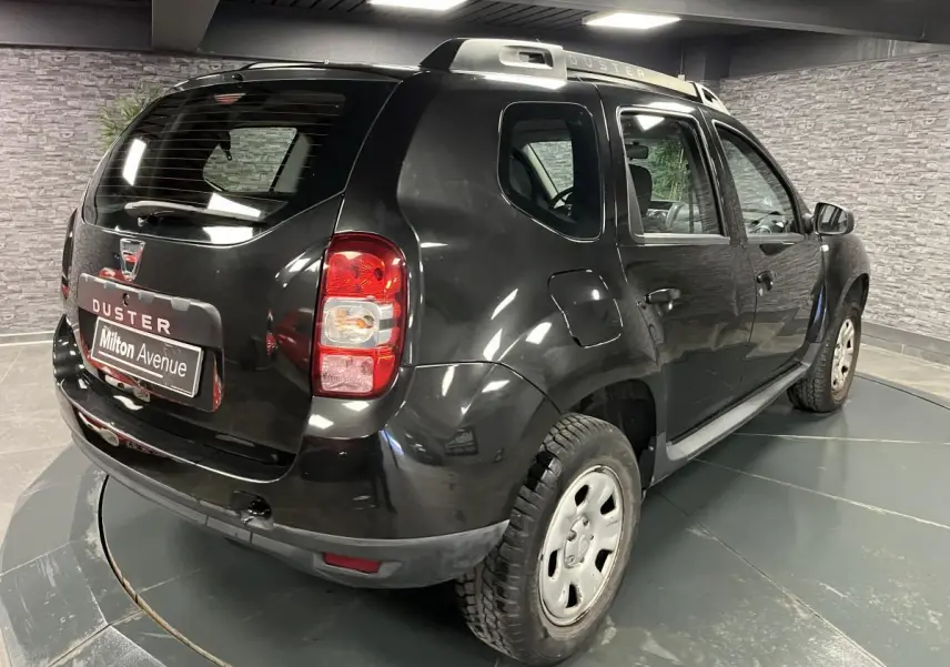 Vue 3/4 arrière droite d'un Dacia Duster noir avec barres de toit et jantes acier en intérieur showroom.