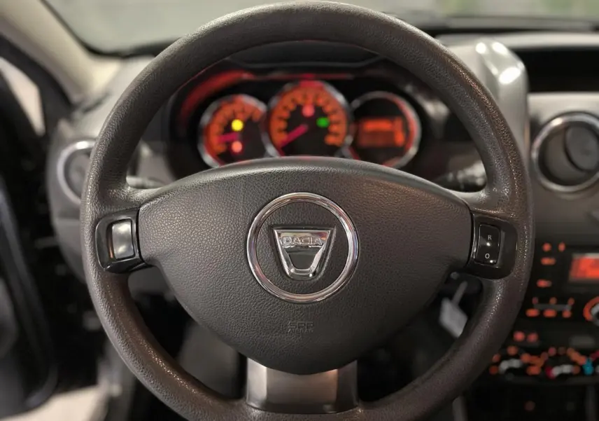 Volant noir avec logo chromé Dacia au centre, tableau de bord éclairé en orange flou en arrière-plan.