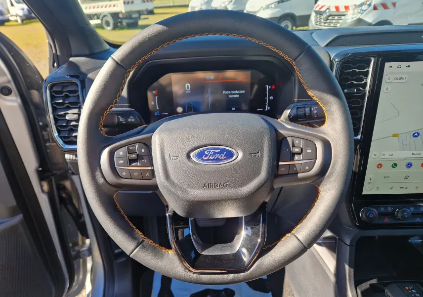Vue rapprochée du volant en cuir noir avec surpiqûres orange du Ford Ranger Super Cabine gris Iconic, tableau de bord et écran tactile visibles.
