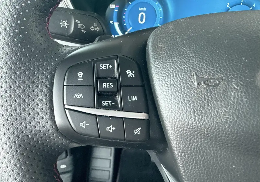 Gros plan sur le volant noir perforé avec commandes de régulateur et volume du Ford Kuga bleu azur, tableau de bord numérique visible.