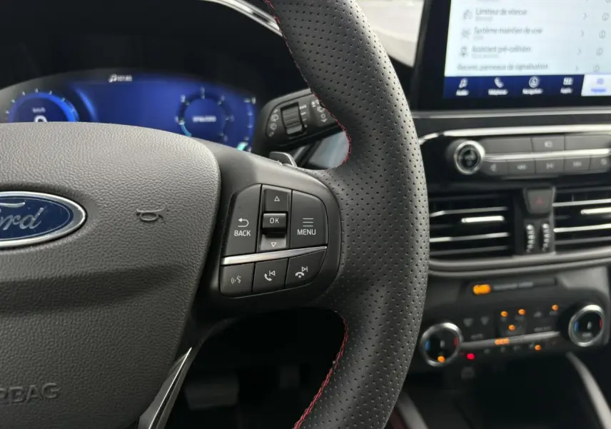 Gros plan sur le volant perforé noir avec surpiqûres rouges et tableau de bord numérique bleu du Ford Kuga 2023.